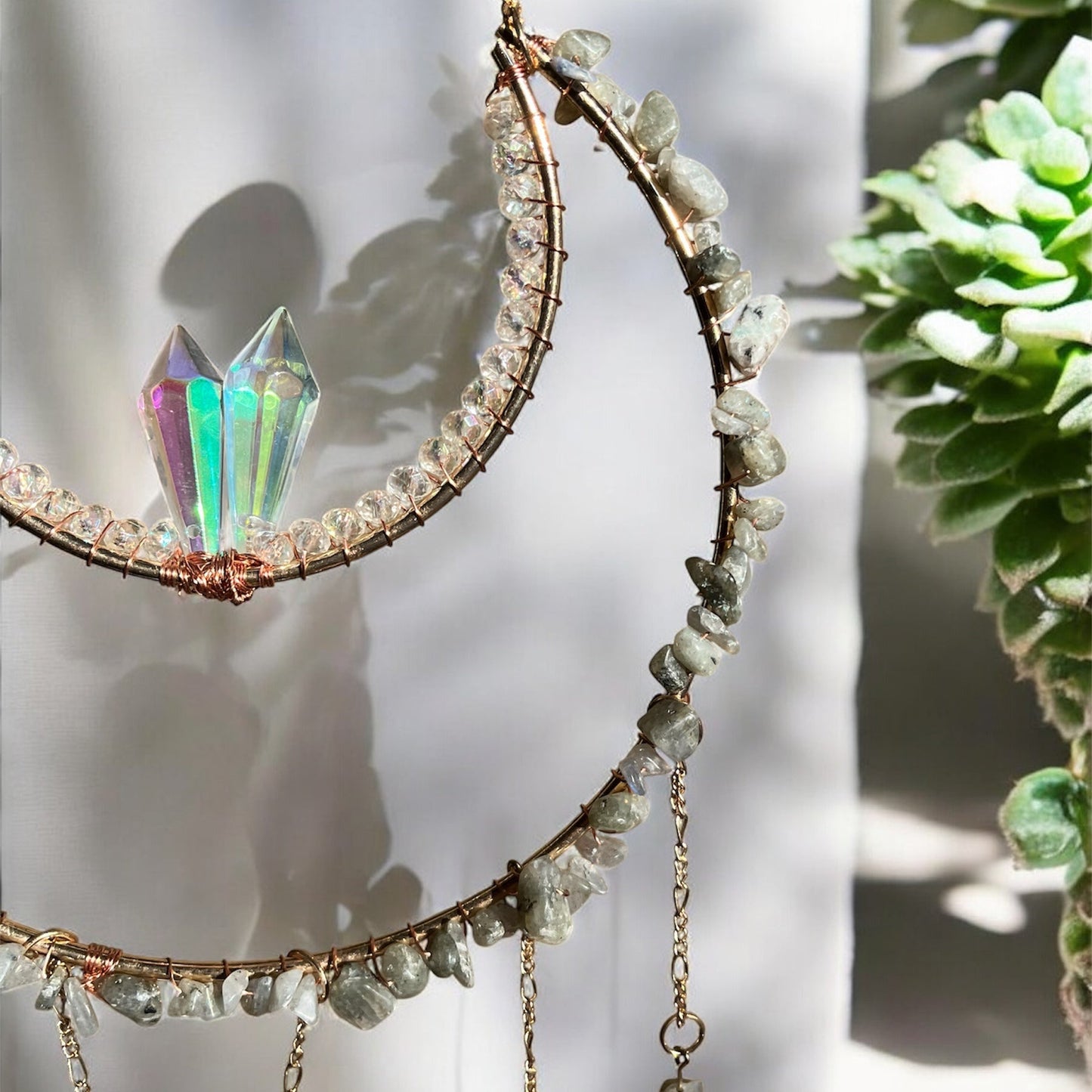 A bohemian-style wall hanging featuring a golden crescent moon frame adorned with small, rough-cut gray and clear crystals, with two pointed iridescent crystals at the top center of the crescent. Below the crescent, five chains hang down, each embellished with various shapes of polished and rough-cut crystals in shades of brown, amber, and iridescent clear, along with small gray and clear beads. A small golden crescent charm also hangs from one of the chains.