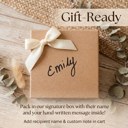 A kraft brown gift box tied with a cream satin ribbon and printed with the name 'Emily' in black script, shown on a rustic wood surface with decorative leaves and the words 'Gift-Ready' at the top.