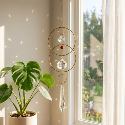Decorative hanging crystal with gold rings and a red gemstone by a window with a plant in the foreground.