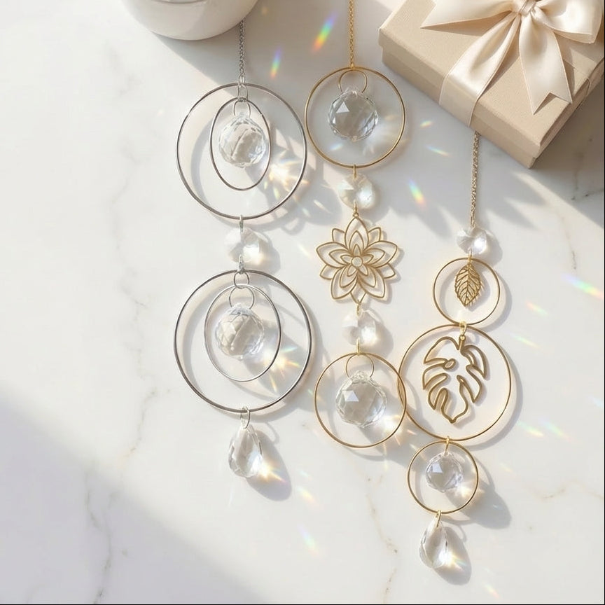Gold-toned suncatcher ornaments with clear glass teardrop beads, pearl-like beads, and cutout floral and leaf designs, arranged on a white marble surface beside a small potted succulent and a cream gift box.