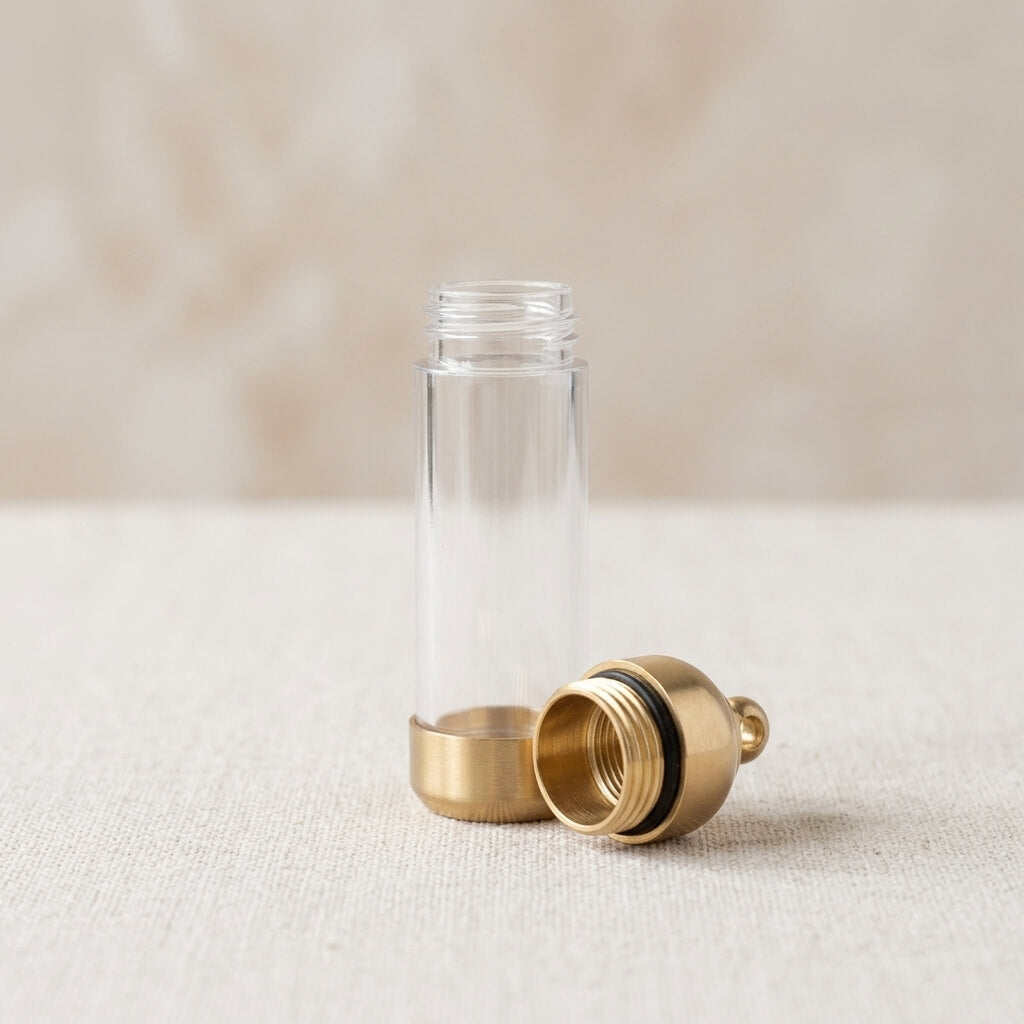 A clear cylindrical bottle with a gold-toned metal base and a matching gold-toned screw-on cap placed beside it on a light beige textured surface.