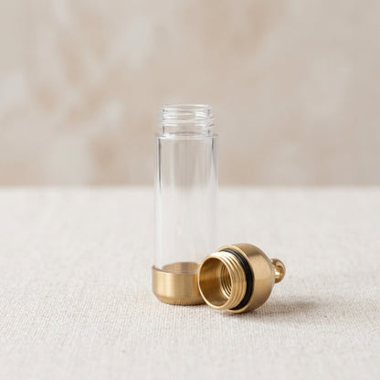 A clear cylindrical bottle with a gold-toned metal base and a matching gold-toned screw-on cap placed beside it on a light beige textured surface.