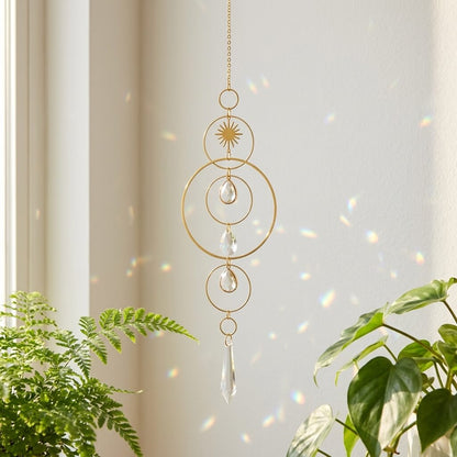 A gold-toned metal suncatcher with clear glass teardrop charms hangs in a sunlit room near a window, with green plants visible at the bottom left and right and rainbow light reflections on the wall.