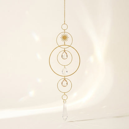 A gold-toned metal suncatcher with concentric rings, a sunburst detail, and clear teardrop and teardrop-shaped crystal prisms, hanging against a warm, softly lit cream background.