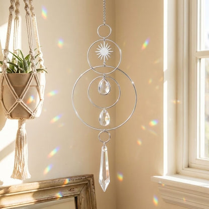 A silver metal suncatcher with concentric rings, a starburst sun face, teardrop crystal drops, and a hanging crystal prism, shown hanging in a sunlit room next to a window with rainbow light reflections on the wall.