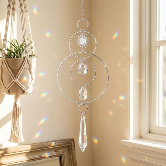A silver metal suncatcher with concentric rings, a starburst sun face, teardrop crystal drops, and a hanging crystal prism, shown hanging in a sunlit room next to a window with rainbow light reflections on the wall.