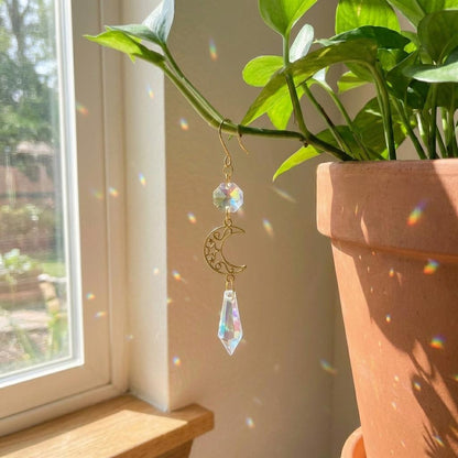 Gold earring with crystal and moon design in front of a potted plant by a window