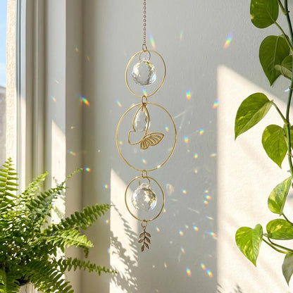 Decorative hanging crystal with gold loops and leaf charms in front of a window with plants.