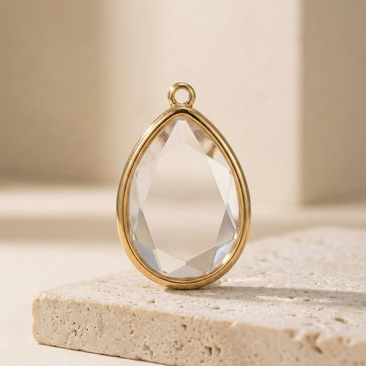Teardrop-shaped crystal pendant with gold frame on a beige stone surface